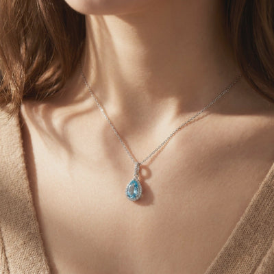 Model wearing a silver necklace with a pear-shaped light blue aquamarine pendant surrounded by a sparkling halo.