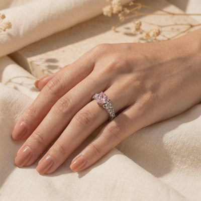 The Rosalique Ring, a solid gold ring with a heart-shaped pink simulated diamond centerpiece and floral band detailing, worn on a hand.