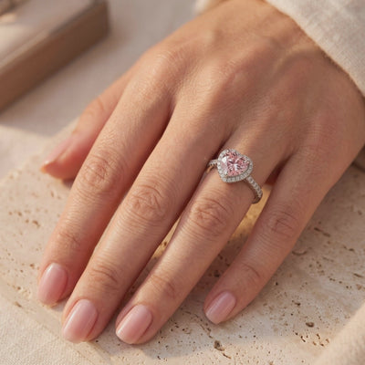 Silver ring with a heart-shaped pink center and sparkling halo, worn on a hand over a textured beige surface.
