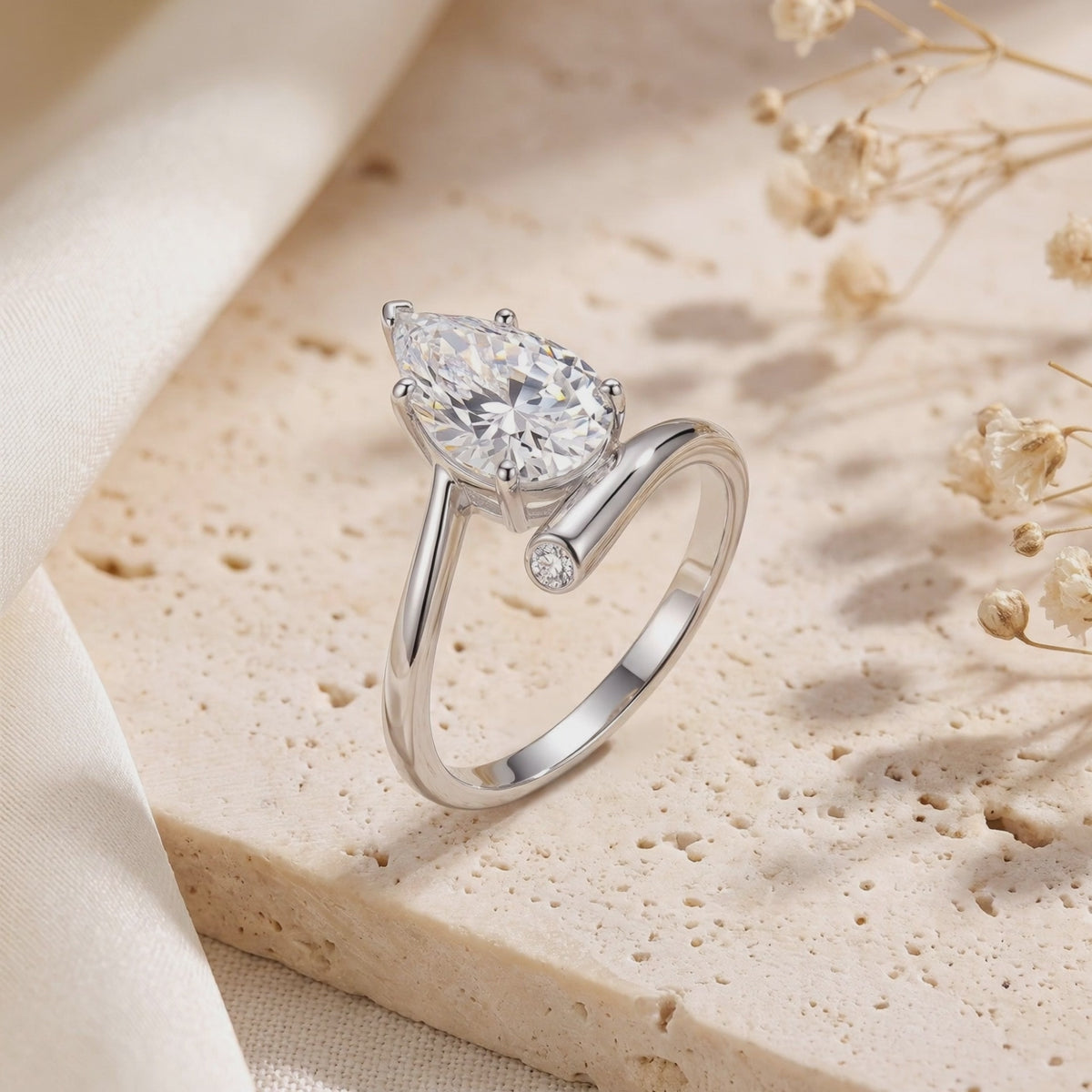 Close-up of a silver bypass ring with a pear-cut simulated diamond and round bezel-set accent on a textured stone block.