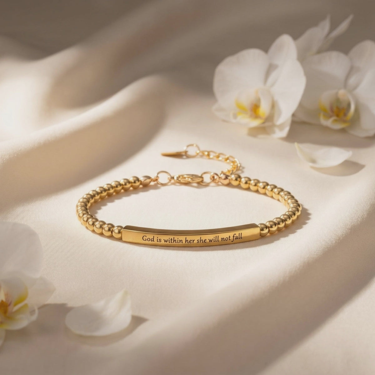 Gold-toned beaded bar bracelet engraved “God is within her she will not fall,” laid flat beside white orchid flowers.