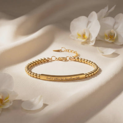 Gold-toned beaded bar bracelet engraved “God is within her she will not fall,” laid flat beside white orchid flowers.