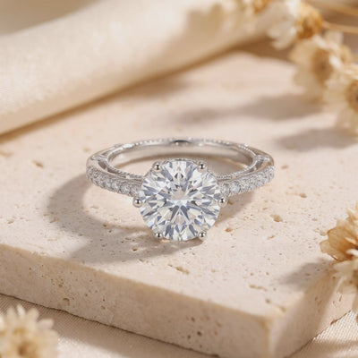 Silver solitaire ring displayed on a light stone block, with a round clear stone, six-prong setting, and pavé accents.